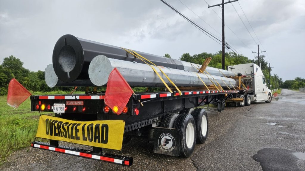 Extendable flatbed truck transporting galvanized steel poles with oversized load signage on a rural road under cloudy skies.