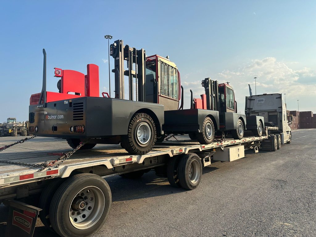 Low-profile step deck truck carrying two large Bulmor side loaders securely fastened, with a setting sun in the background.