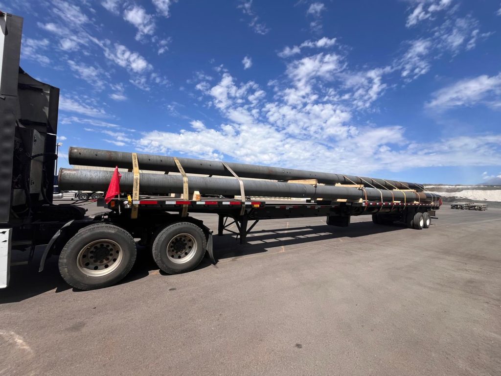 Stretch flatbed truck carrying four large poles securely strapped for transport under a clear blue sky.