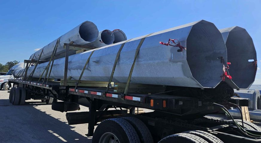Stretch flatbed trailer carrying oversized metal poles secured with straps, ready for transportation under a clear blue sky.