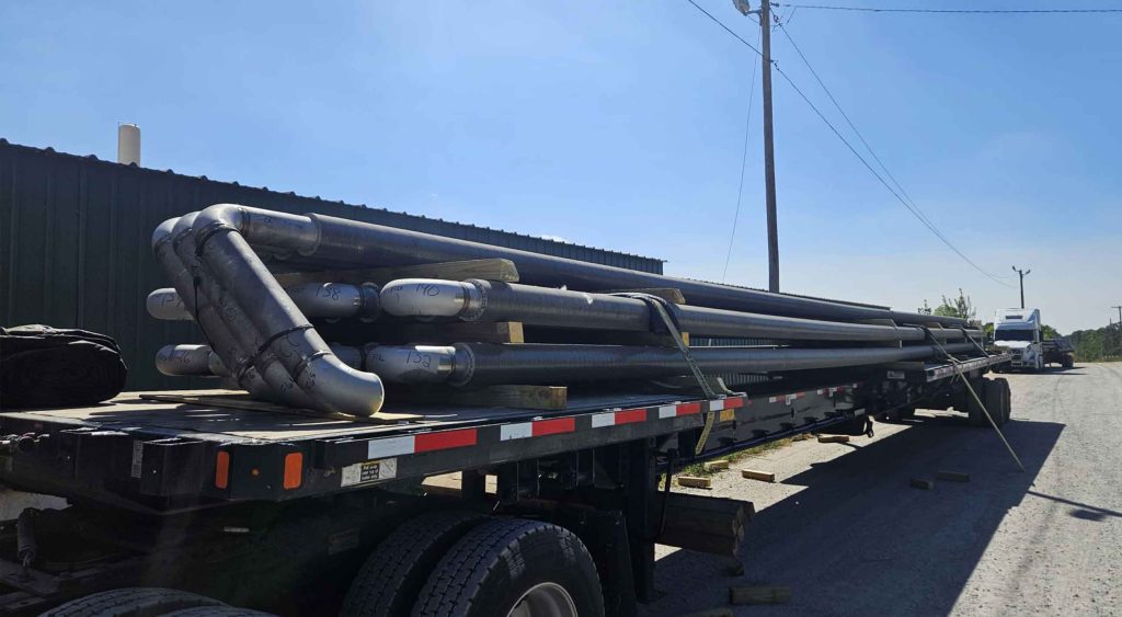 Flatbed trailer carrying large industrial pipes secured with straps, ready for transport under clear blue skies.