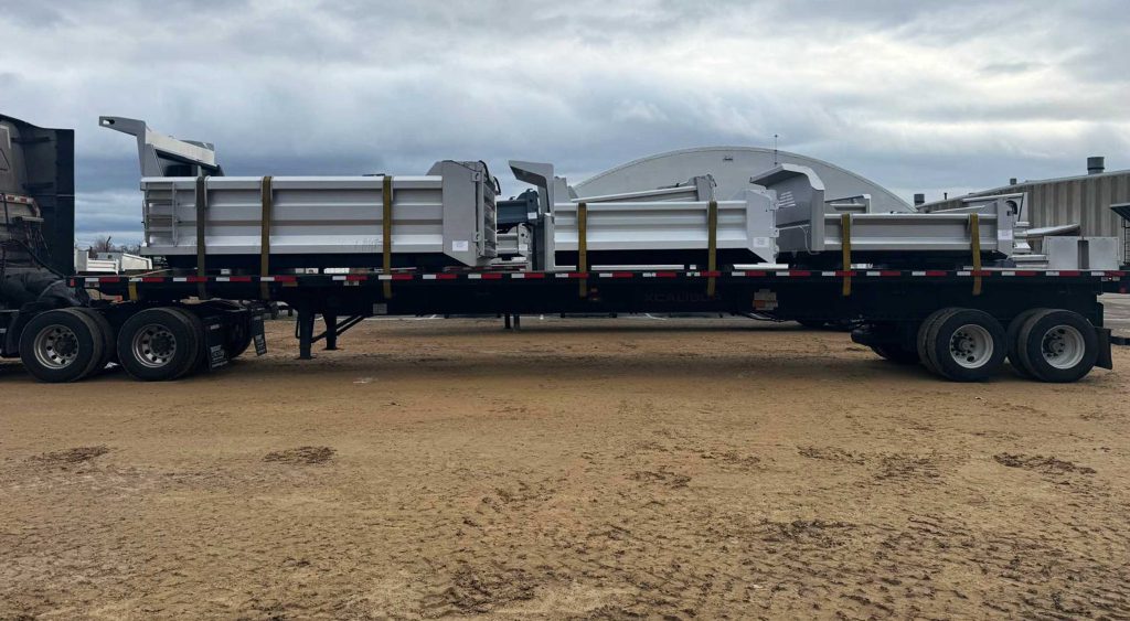 Flatbed trailer carrying truck bodies and hoists, securely strapped, ready for delivery under a cloudy sky.