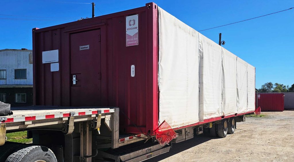 Step deck trailer carrying a red mobile office/storage unit secured with straps, ready for delivery under a clear blue sky.