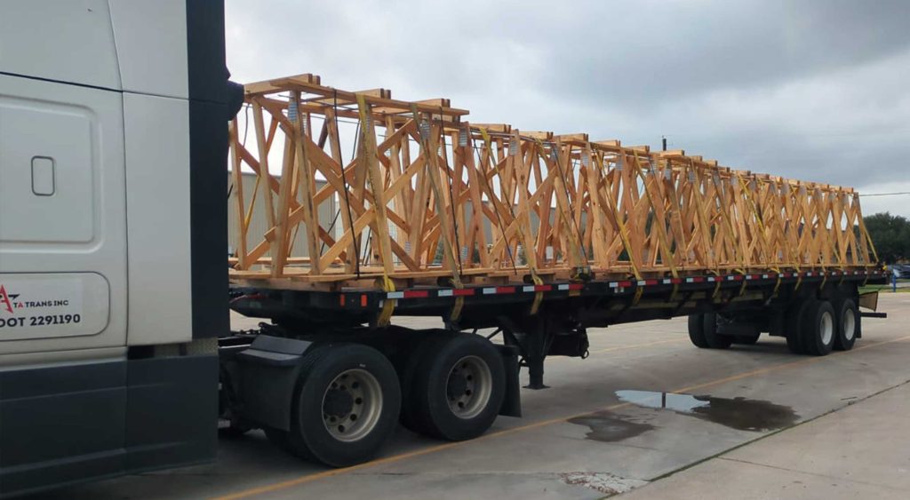 Flatbed trailer loaded with wooden A-frames for construction, securely strapped, parked at a delivery location under cloudy skies.