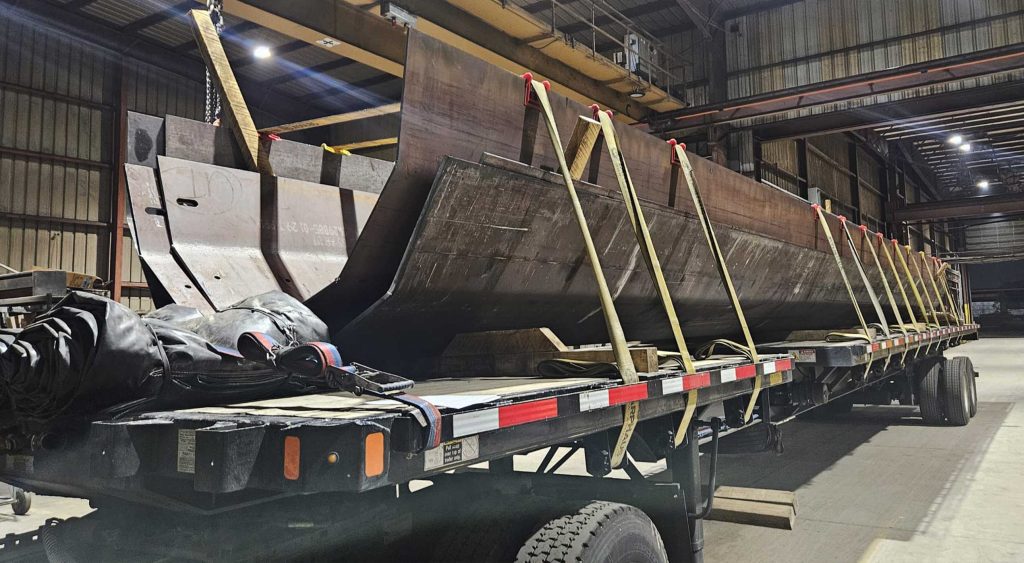 Extendable Flatbed trailer loaded with heavy steel beams secured by multiple straps inside a warehouse, ready for transportation.