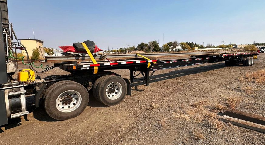 Extendable Flatbed trailer carrying an oversized industrial load securely strapped, photographed on a sunny day at a logistics hub.