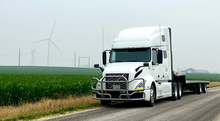 White semi-truck with a flatbed trailer representing the future of freight in 2025 on a rural road with wind turbines in the background.