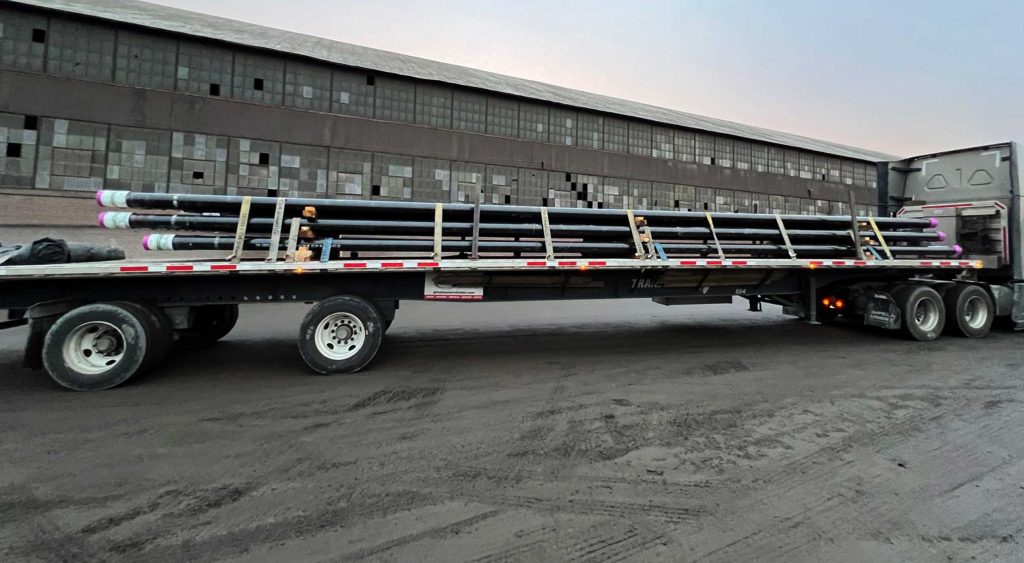 A flatbed truck loaded with 32 large tubing pipes, securely strapped and ready for transport. The truck is parked in front of an industrial building under a cloudy sky, showcasing precision in heavy freight handling.
