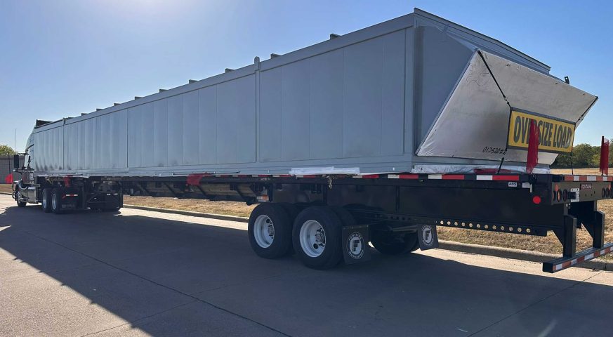 A stretch flatbed truck carrying an oversized upper enclosure assembly under clear blue skies, ready for transportation from Denison, TX, to Phoenix, AZ.