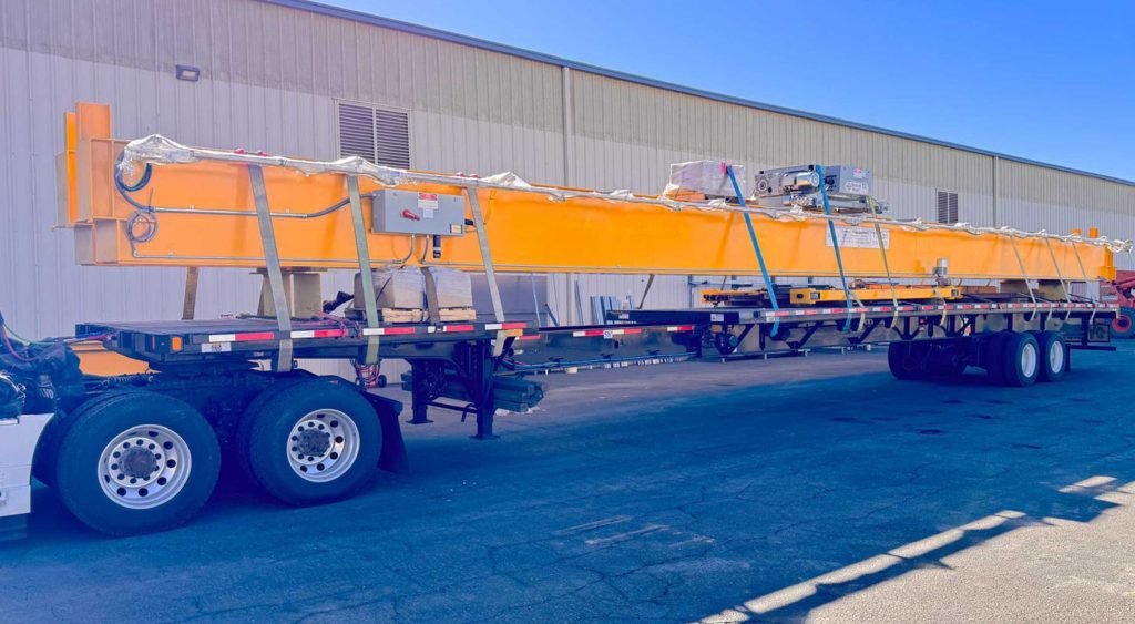 A stretch flatbed truck loaded with a large, bright yellow crane component, parked outside a warehouse under a clear blue sky.