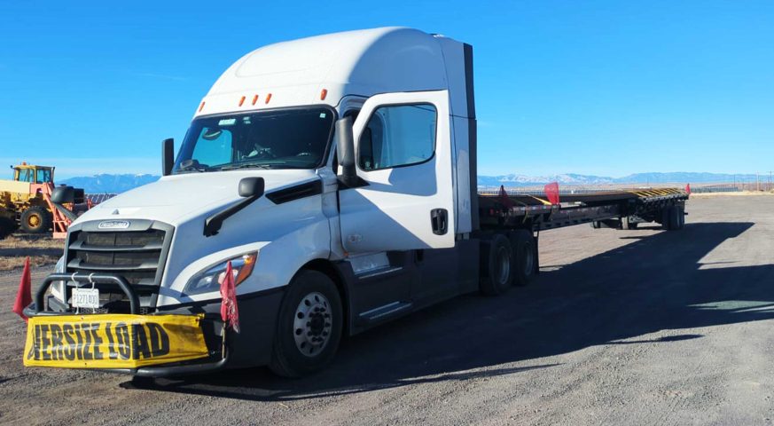 Freightliner truck carrying an oversized 136RE T-rail assembly, securely fastened for transport at a logistics yard under a clear blue sky.
