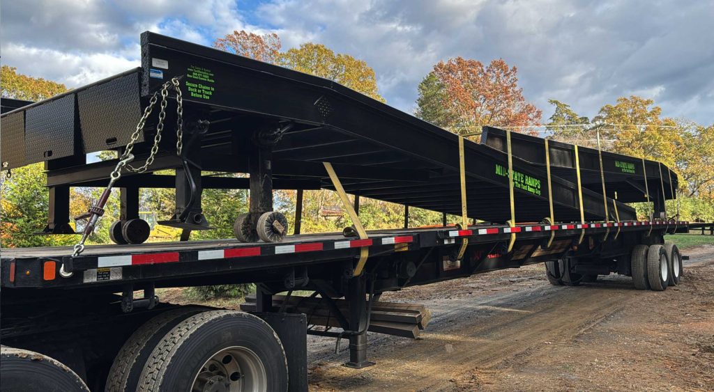 A flatbed truck loaded with a black mobile all-purpose yard ramp, securely fastened with straps and ready for transport. The ramp features detailed structural components and is set against a background of trees and an overcast sky.