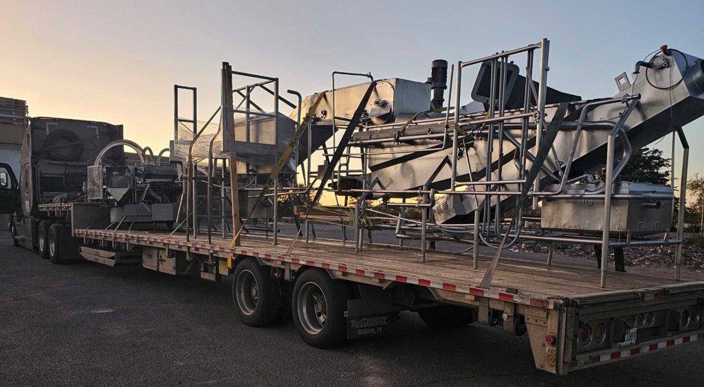 A flatbed trailer carrying large stainless steel dairy processing equipment, securely loaded for transport at sunset.