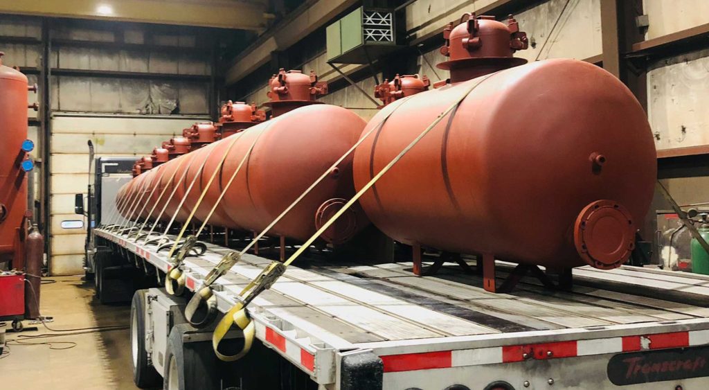 A flatbed trailer loaded with multiple red steel tanks, securely strapped inside a warehouse, ready for transport.