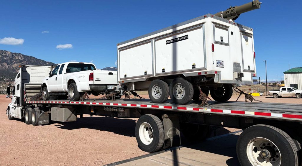 A flatbed trailer loaded with a white pickup truck and a snow control trailer, secured for transport under a clear blue sky.