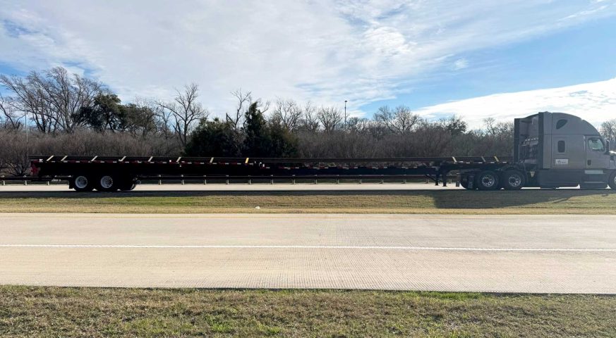 A TA Trans INC stretch flatbed trailer transporting long rail sections on a highway, ensuring safe and efficient oversized freight delivery.