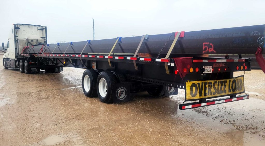 A stretch flatbed trailer transporting 65-foot steel beams securely fastened for long-distance delivery under overcast skies.