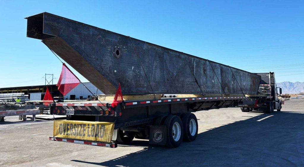A stretch flatbed trailer transporting a massive box girder with red safety flags and an 'Oversize Load' sign under a clear sky. extendable flatbed hauling bridge box girder for construction project