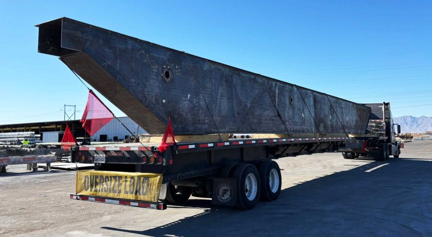 A stretch flatbed trailer transporting a massive box girder with red safety flags and an 'Oversize Load' sign under a clear sky. extendable flatbed hauling bridge box girder for construction project