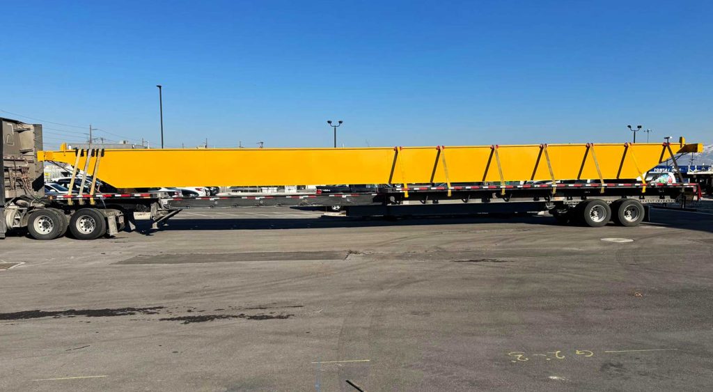 A stretch flatbed trailer carrying a large yellow crane girder, securely fastened for transport in an industrial yard.
stretch flatbed carrying crane girder for jobsite delivery