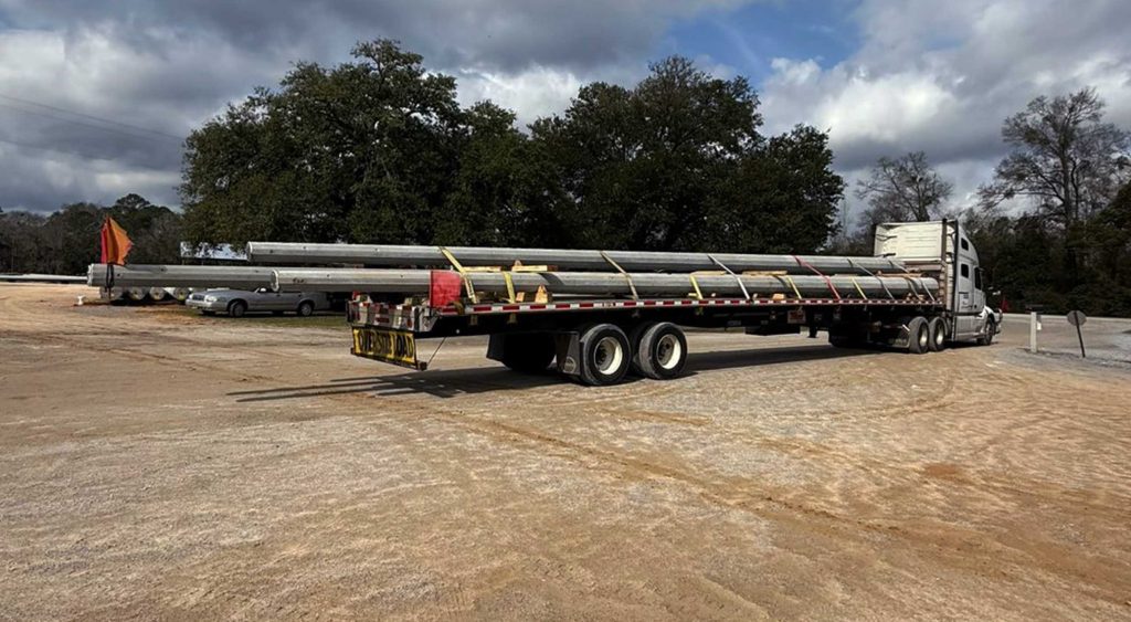 Flatbed trailer hauling long utility poles secured with yellow straps and flags at rear for oversize load transport.