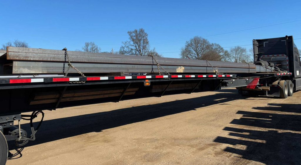 Flatbed truck loaded with long steel beams secured for delivery on a clear day in Alabama.