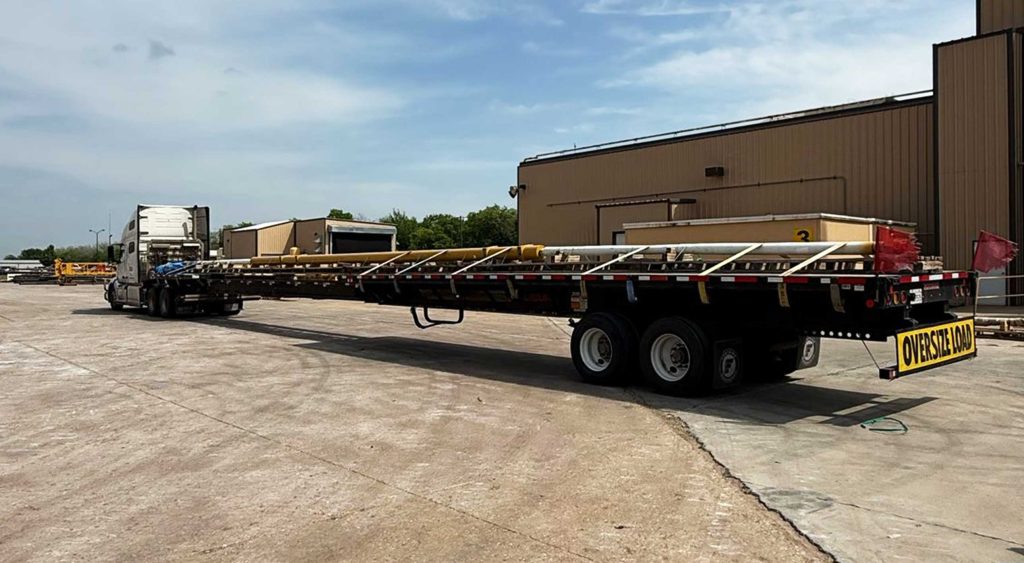 Flatbed truck with stretch trailer carrying oversized hydraulic cylinder marked with “Oversize Load” sign in front of an industrial facility.