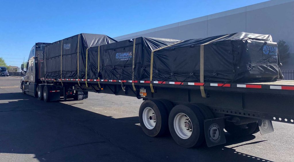 Flatbed truck loaded with fully tarped mid freezer panels, parked in front of a warehouse under clear blue skies. The load consists of 7 pallets, tightly secured for temperature-sensitive delivery from Arizona to Idaho.