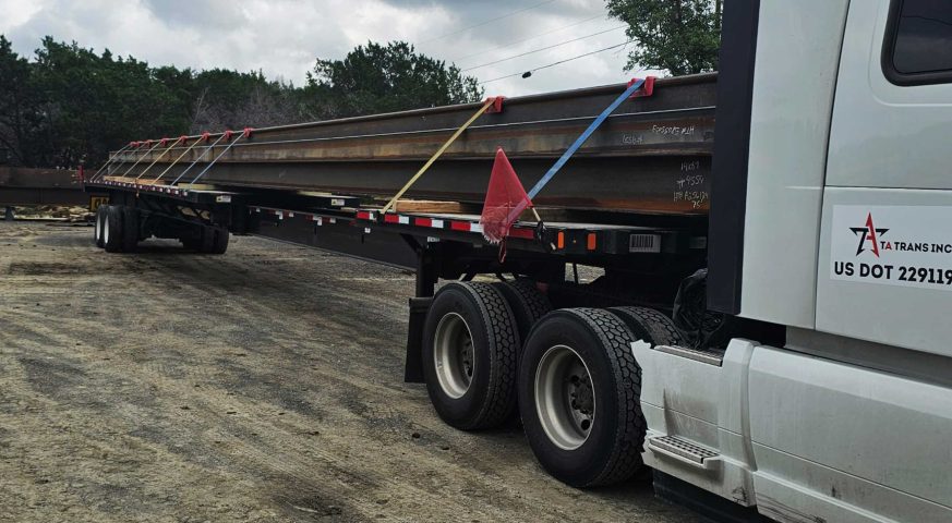 A stretch flatbed trailer loaded with long steel H-pile beams, secured with ratchet straps and flagged for oversize load, parked at a jobsite. TA Trans Inc. truck in view.