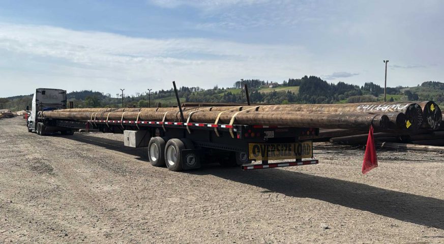 A stretch flatbed trailer loaded with six 100-foot utility poles, secured and ready for transport at a lumber yard in Sheridan, Oregon. The trailer is marked for an oversize load, parked on gravel with a scenic background of trees and hills.