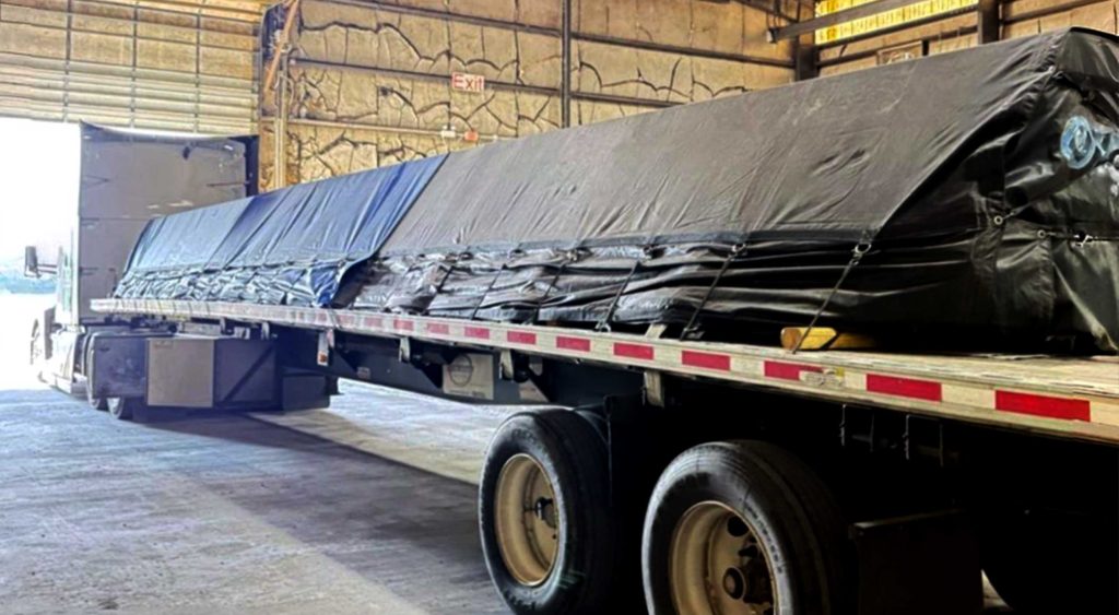 Flatbed truck hauling steel bars with tarps en route from Mississippi to Nebraska.