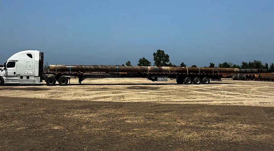 White semi-truck hauling 90-foot steel pipe on a stretch flatbed trailer in a dirt yard under a clear blue sky.