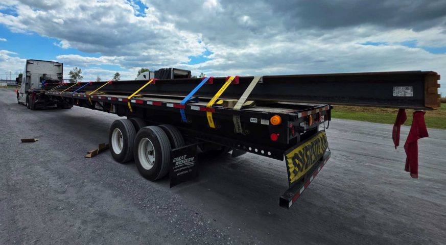 Stretch Flatbed trailer hauling long steel beams, tightly strapped with chains and tarps, flagged for secure transport. extendable flatbed transporting long steel beams