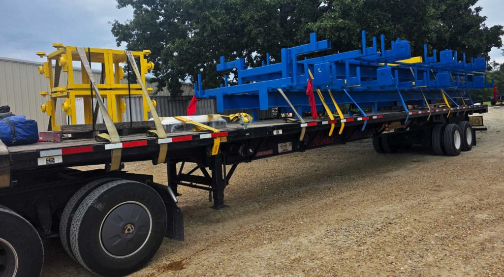 Flatbed trailer carrying blue and yellow steel catwalk and tower components, securely strapped with red and yellow flagging.