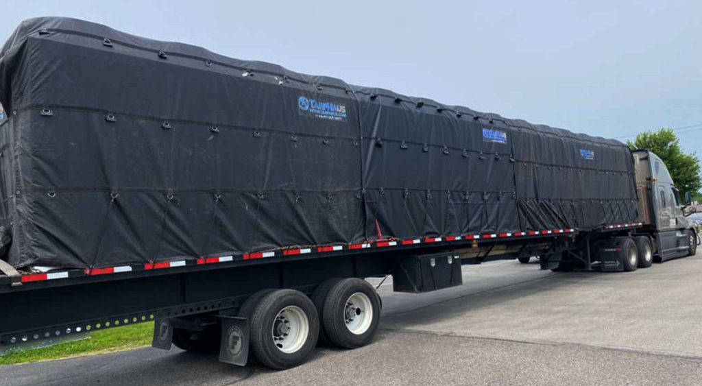 Extendable flatbed trailer hauling an oversized 57 ft industrial chiller, fully tarped and secured.