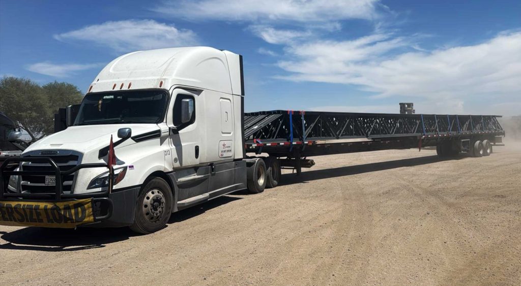 Oversize stretch trailer loaded with long steel trusses in a dusty lot.