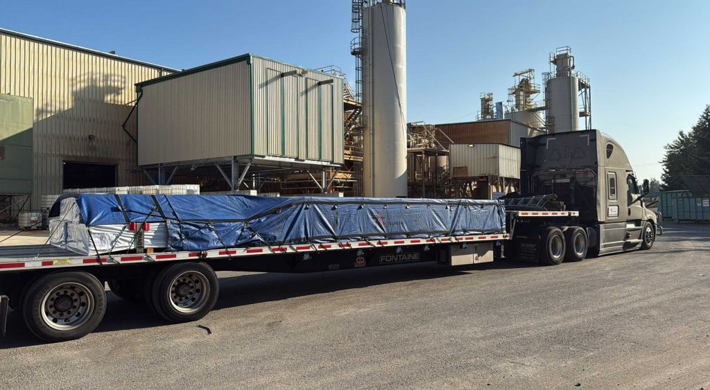Flatbed loaded with tarped pallets of siding outside a manufacturing facility.