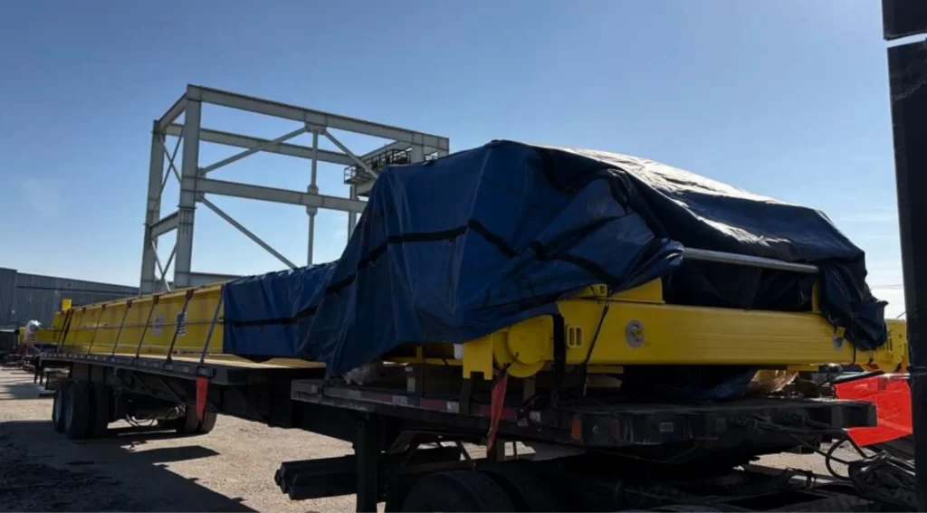 Yellow crane component tarped and secured on a flatbed trailer at a loading site under daylight.