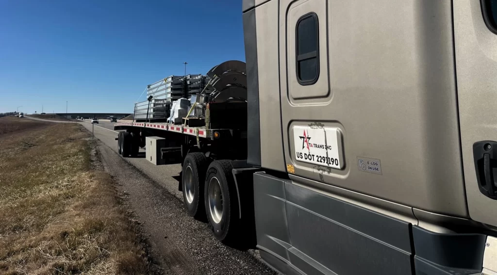 Light-weight electrical conduit cargo strapped on a flatbed trailer, roadside under clear blue skies.