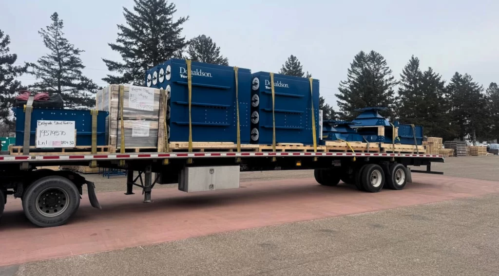 components secured on a flatbed trailer, ready for delivery in a yard with pine trees in the background.