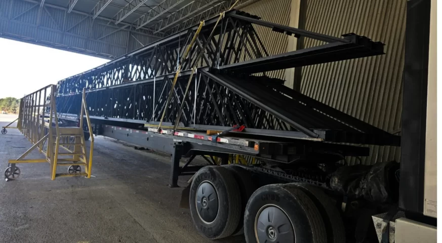 Flatbed trailer hauling stacked 82-ft steel joists inside a loading facility.