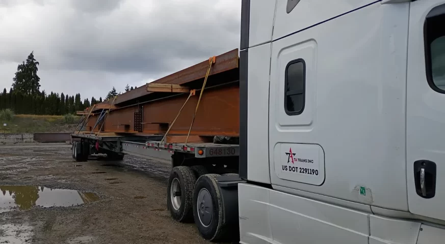 Stretch Flatbed truck hauling large steel beams secured with straps at a job site, representing high construction demand driving flatbed rates up in the U.S. in 2026.