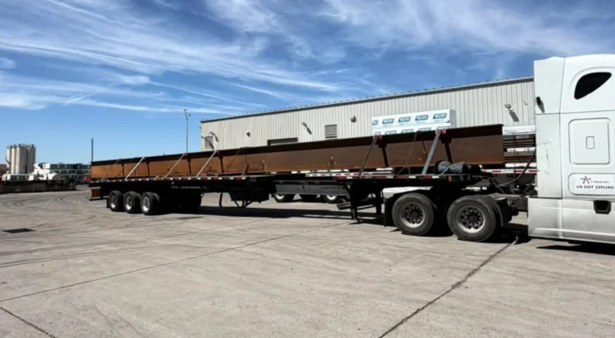 Side view of a TA Trans truck and trailer carrying long steel beams in an industrial yard.